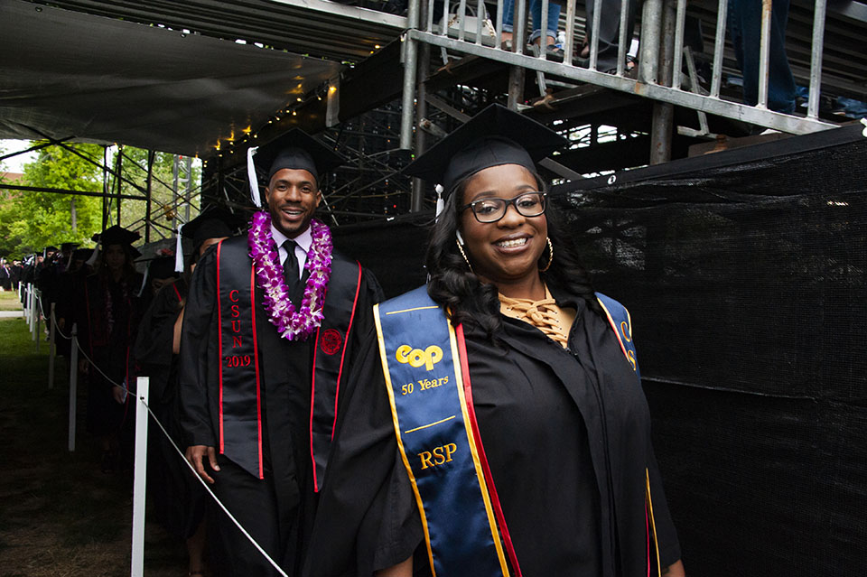 Smiling college student is wearing a navy blue sash that says 'EOP 50 Years RSP' and arriving to a graduation ceremony.
