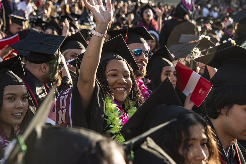 A female student in a sea of seated graduates waves to loved ones.