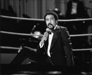 Richard Pryor sitting in a boxing ring and speaking into a microphone. Photo by Guy Crowder | Tom & Ethel Bradley Center.