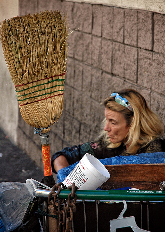 A homeless woman crouches on the ground with her belongings in a shopping cart.