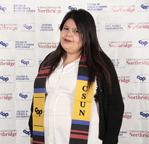 A smiling woman with dark brown hair sporting a CSUN graduation sash, white blouse and black cardigan.