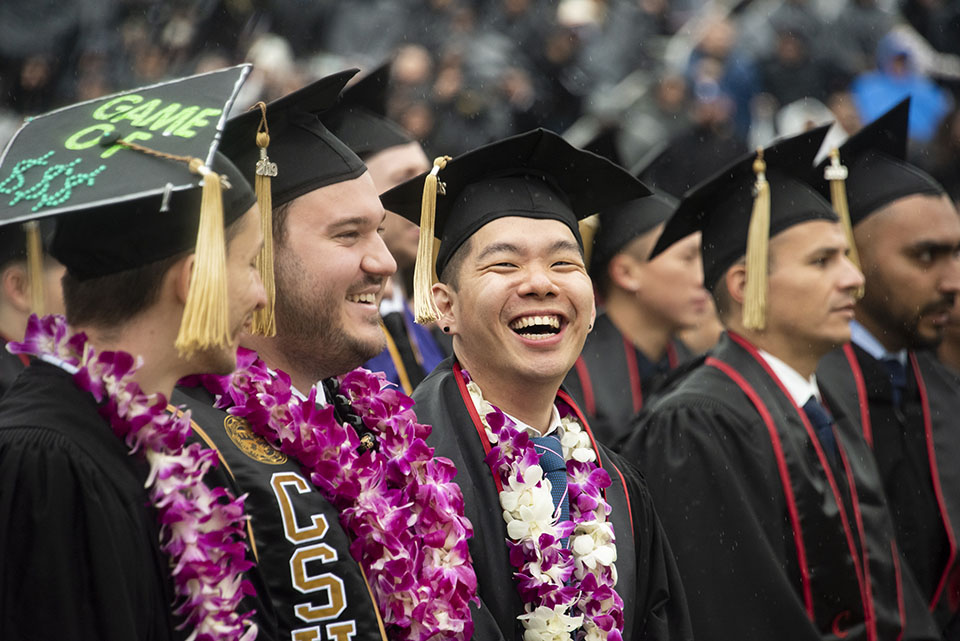 Three male students in a graduation cap and gown smile at their commencement ceremony.