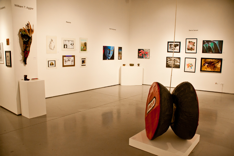 Fifteen pictures hanging on two walls in the art gallery. A large statue replica of a yo-yo sits in the foreground. All pieces were selected by art teachers from their respective high schools.