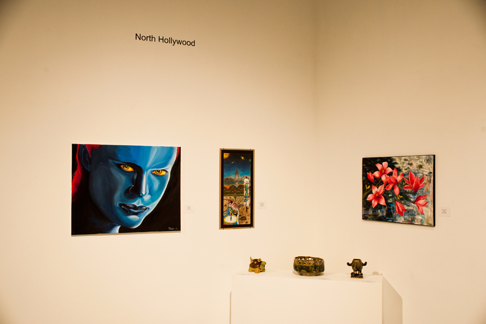 Three paintings and three small sculptures by students at North Hollywood High School. A large painting on the left-hand side depicts a blue female face with fiery red hair and yellow eyes. The piece is entitled “Fatal Beauty,” an oil-on-canvas, by Talia Dutton. An oil painting on the right-hand wall depicting a floral scene is untitled by Valeriana Stefan.