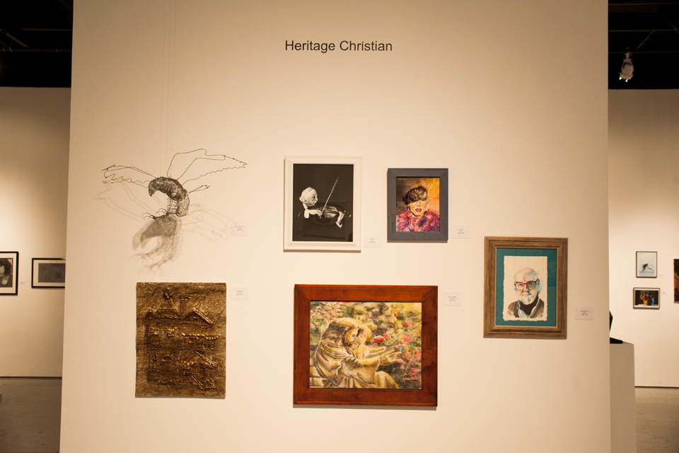 A wall in the gallery where six student works from Heritage Christian School of Northridge are featured. This display includes a wire sculpture of a bird, four portraits—three male and one female—a golden mixed media piece that includes a house-like shape.
