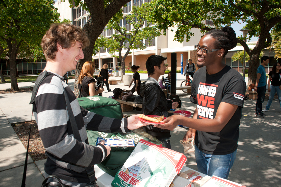 A male student gives a slice of pizza to another student.