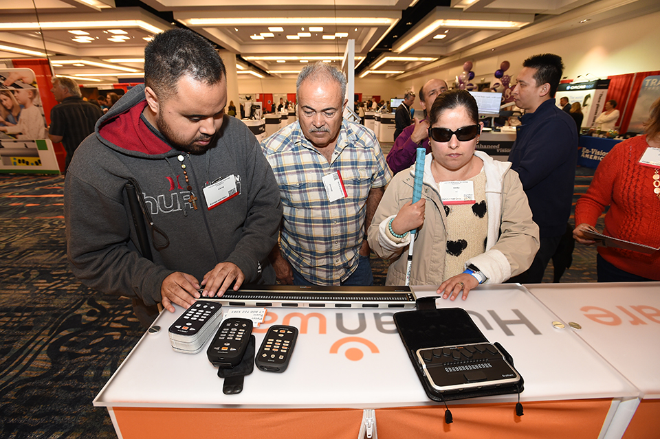 Three individuals with vision impairments inspect braille products, electronic magnifiers, and digital talking book players.