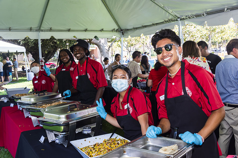 Volunteers wearing red CSUN shirts and black aprons stand, smiling behind a buffet table
