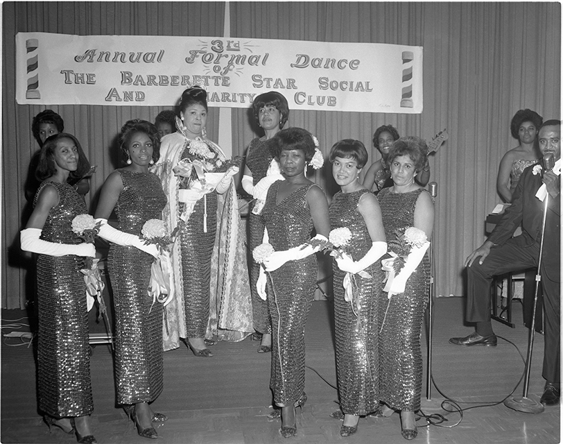 A vintage photo of a group of women onstage at a formal dance of the Barberette Star Social and Charity Club. A man in the far right speaks into a microphone.
