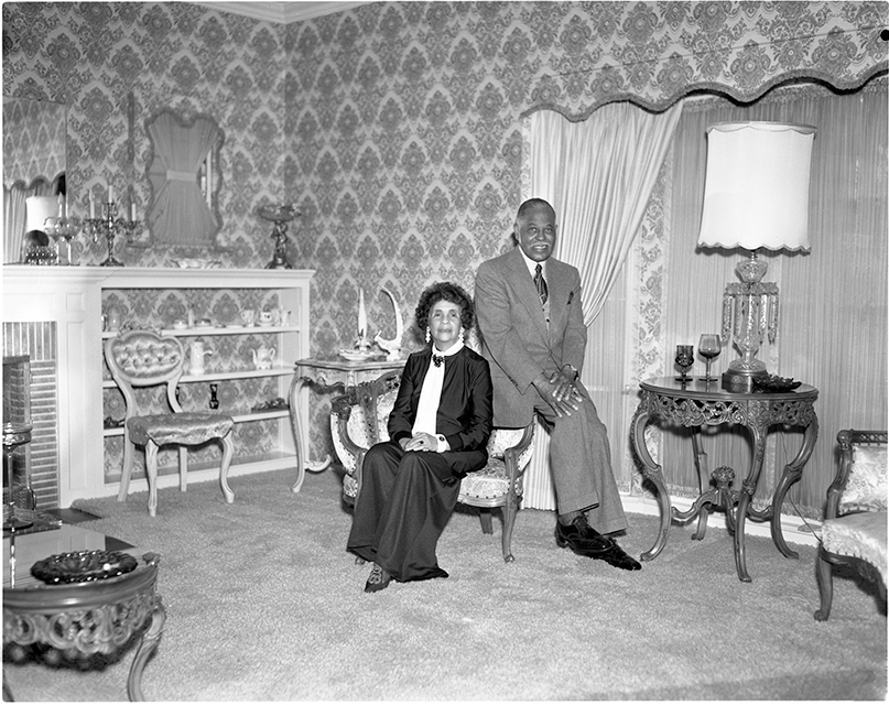 An African American couple, seated, pose for a portrait in a well appointed room inside of a home.