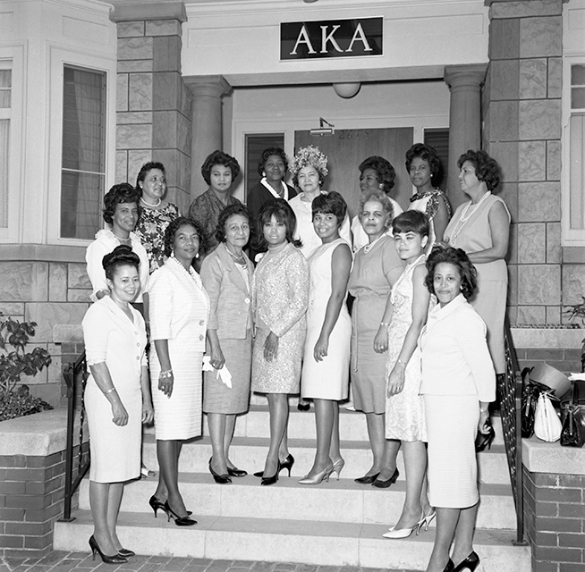 A vintage photo group photo of members of Alpha Gamma Omega, and nine high school graduates who were receiving scholarships, in front of the Alpha Kappa Alpha fraternity house.