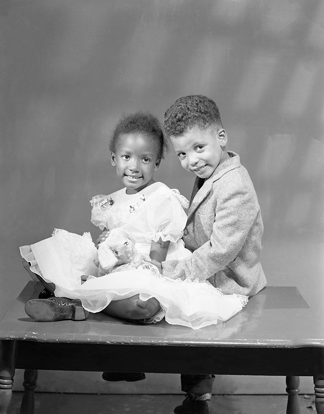 A young African American girl and a young African American boy, both smiling, seated closely to one another, pose for a photographic portrait.