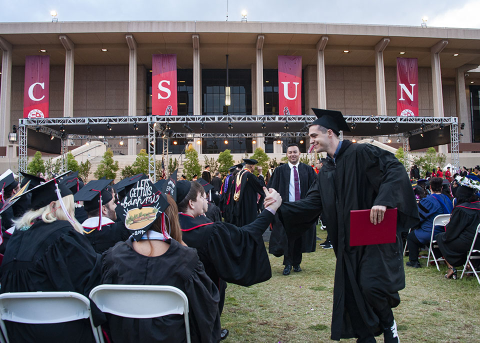 A CSUN graduate holding his diploma shakes the hand of a seated fellow graduate as he exits the stage at 2019 commencement.