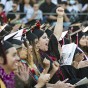 A row of graduates in caps, gowns, sashes and flower leis clap and cheer, at Commencement 2022. One graduate raises her fist in the air, in triumph.