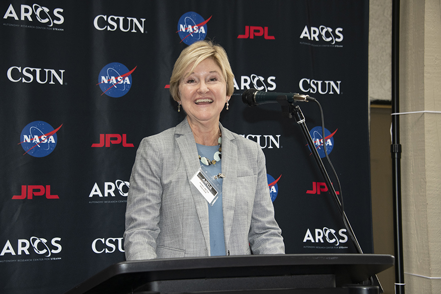 A blonde woman in a gray plaid suit and name tag speaks onstage behind a podium.