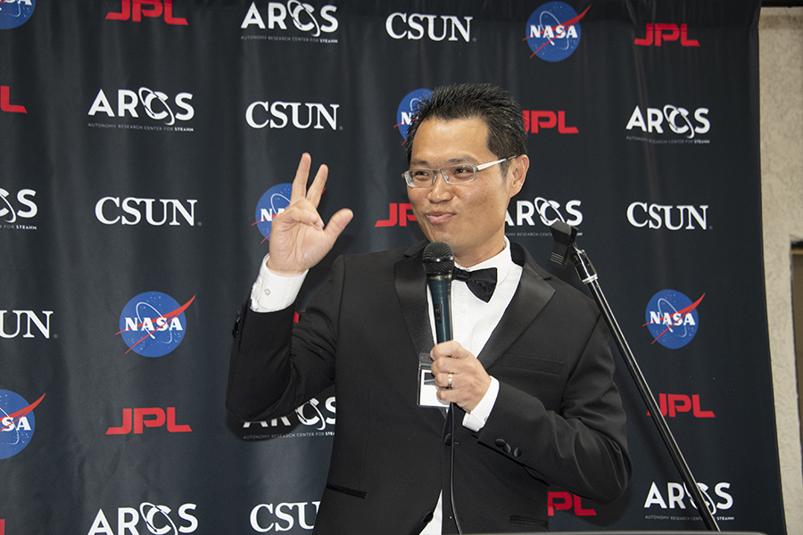 A man in a black tuxedo sports a bowtie and glasses and speaks onstage with a microphone in his hand.