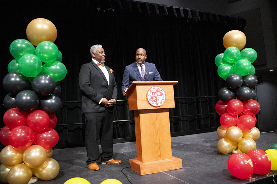 Cedric Hackett and Yan Searcy stand at podium on stage, flanked by colorful balloon towers.