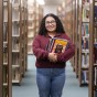 Betsy Benavides stands in an aisle of the University Library stacks, surrounded by tall shelves of books. She stands holding a stack of books.