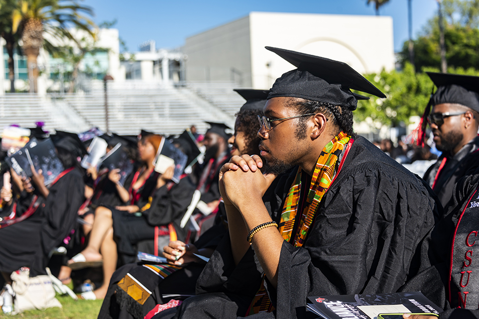 A student in cap and gown in the foreground listens intently while seated in the audience