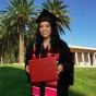 A female student holds a diploma and wears a graduation cap and gown in front of a virtual CSUN library background.