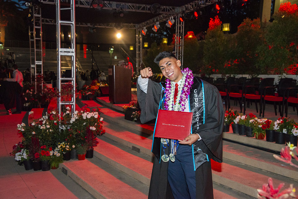 A smiling college graduate is holding a red diploma and pointing at the camera on a stage.
