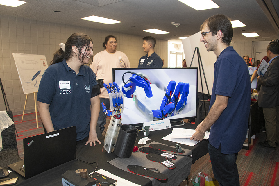 Two students stand next to a prosthetic arm on the table between them.