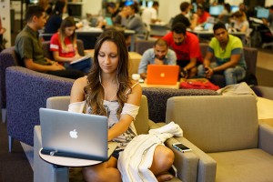 Student sits in foreground with open laptop in front o her. Students in background also working on laptops.