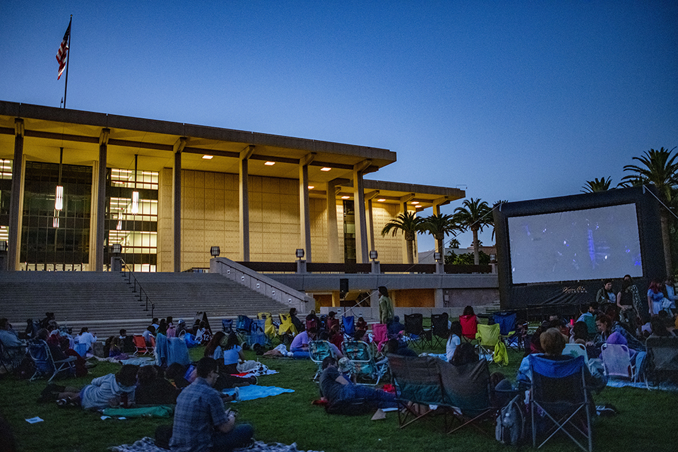 People are seated on the grass in front of a movie screen on the University Library Lawn with the University Library lit up in the background.