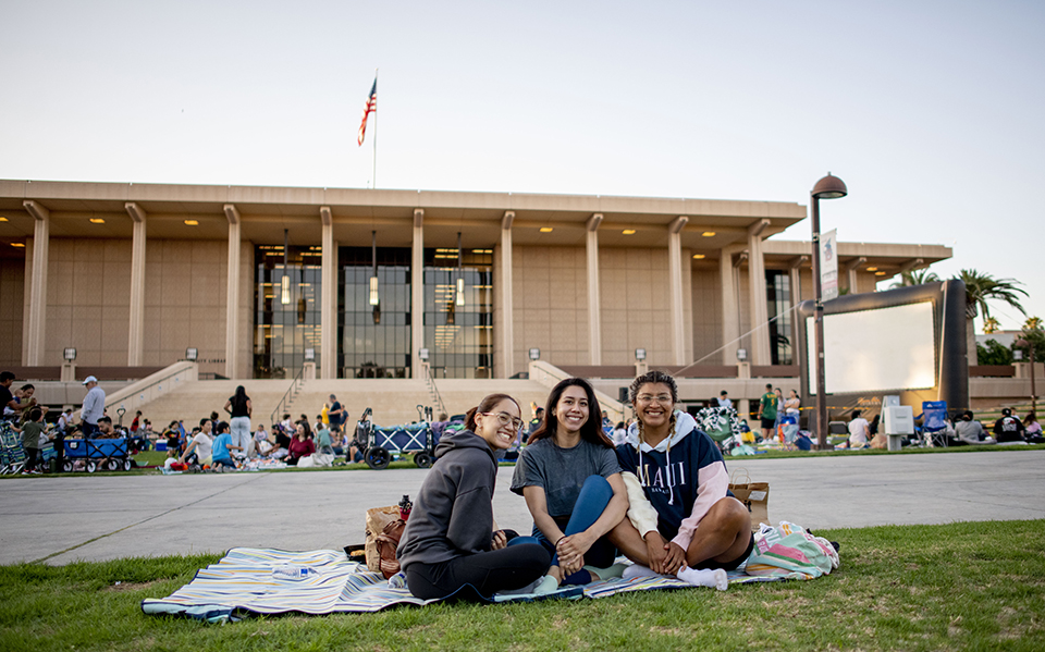 Three young women sit on a blanket on the University Library Lawn, they are smiling at the camera. People behind them are on the grass in front of a movie screen