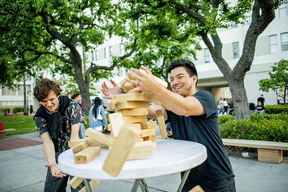 Two young men stand on either side of a collapsing Jenga block tower. One man is reaching out to catch some of the falling blocks.