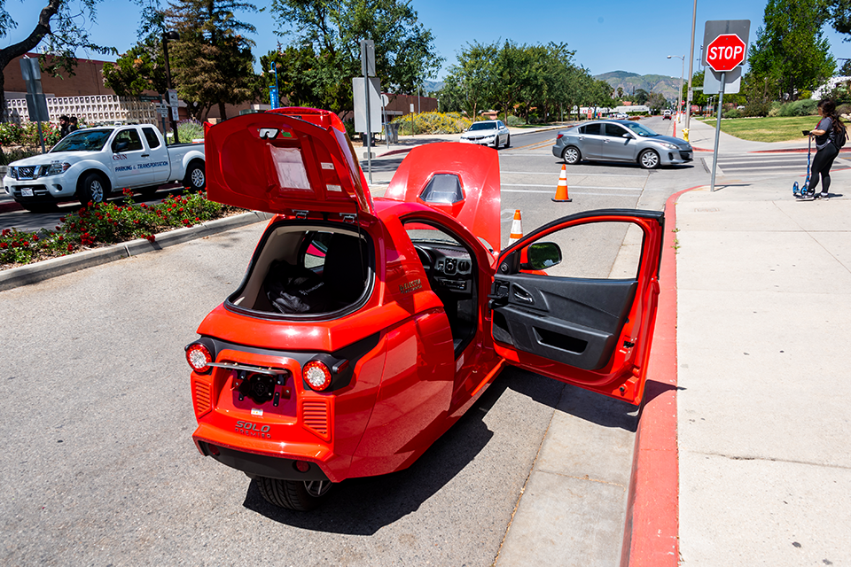 Three wheeled red car in roadway with its trunk, hood and passenger door open