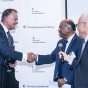 Dean Chandra Subramaniam shakes hands with Rick Caruso, while Fred Gaines stands next to them, holding a microphone.