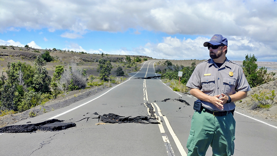 Mike Theune in his National Park Service uniform stands on a road with deep cracks with trees and smoke in the background.