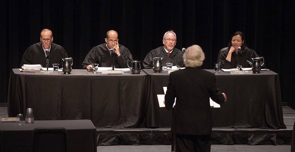Four judges, robed in black, sit at a black cloth-covered table, listening to an attorney present his case. He is standing at a podium.