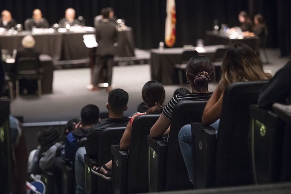 A back view of rows of seated students in the Plaza del Sol Performance Hall, seated and watching the California Court of Appeal hold a live session on stage.