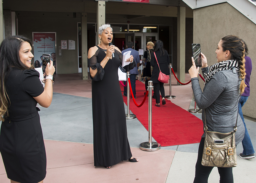 Deborah Wallace speaks into a microphone as two women film her with cameras beside the red carpet.