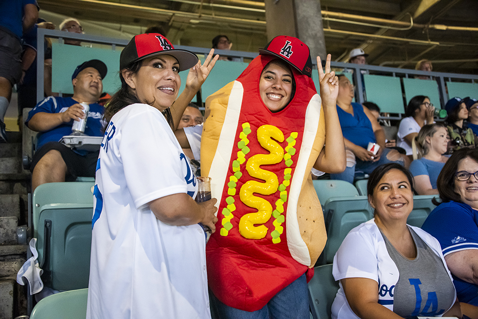A woman dressed as a hot dog in a CSUN-red Dodger hat makes the peace sign while bystanders look at her.
