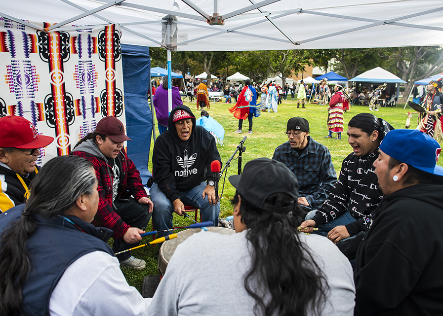 A group of men sing in a circle around a microphone and a drum at the 2019 CSUN Powwow.