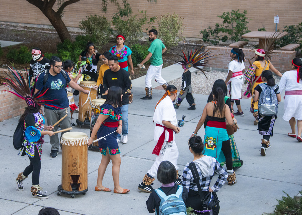 Procession dancers moving in a circle formation with drum players following as several students watch.