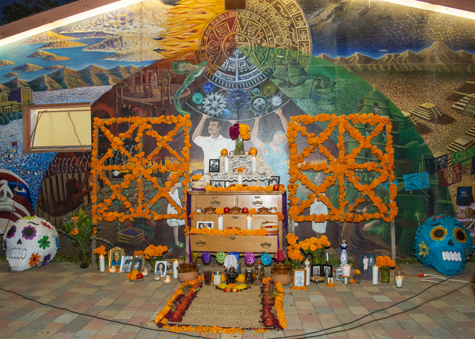 A grand altar with flowers, candles, photos and sugar skulls stands in front of a mural.