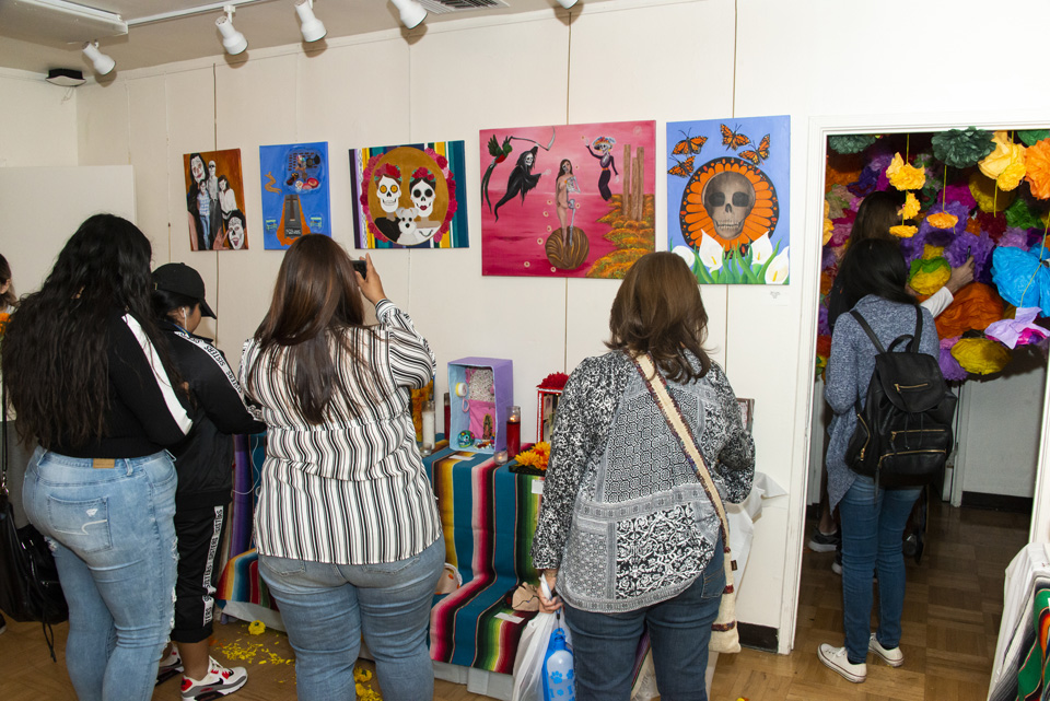 Two women stand in front of a line of art pieces above several altars, one is taking pictures.