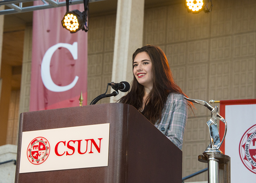 Nicole Maines speaks at a podium in front of the Oviatt Library and CSUN administrators on stage.
