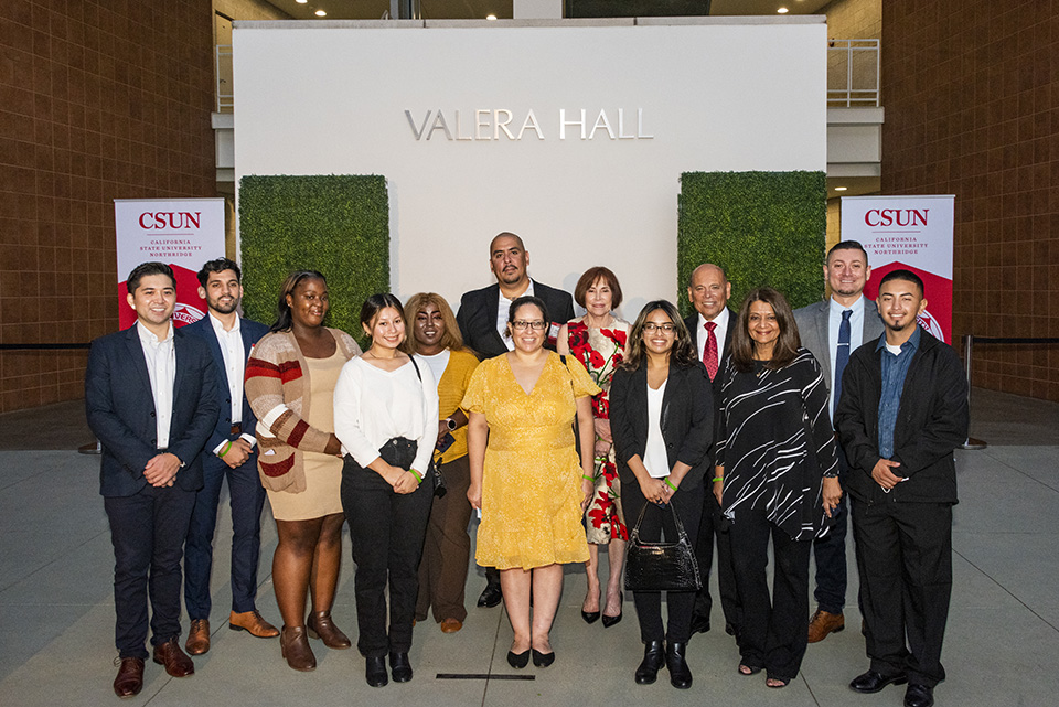 A group of 13 people, of various ages, gender and ethnicities, pose with Milt and Debbie Valera in front to the Valera Hall building signage.