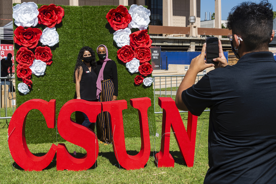 Two people pose behind giant CSUN letters and in front of a flowery backdrop, while another person takes their photo