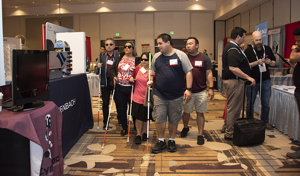 A group of individuals with vision impairments walk through the CSUN Conference exhibit hall. Four of the five carry white canes.