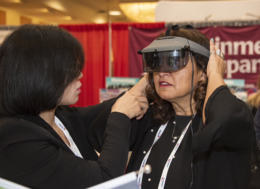 A woman assists another woman in putting on a vision-enhancing headset with dark glasses.