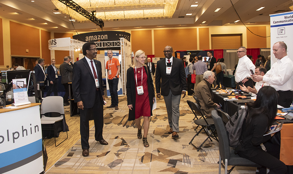 CSUN President Dianne F. Harrison walks with William Watkins and Dwayne Cantrell down a wide aisle of the Assistive Technology Conference exhibit hall.