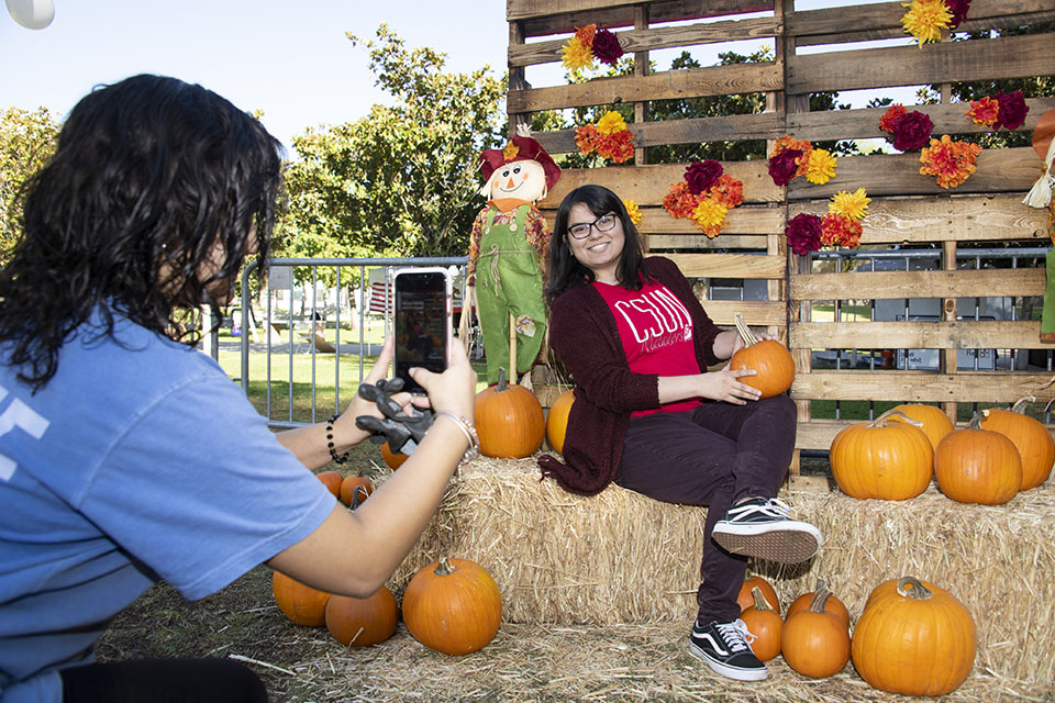A student poses for pictures with her pumpkin in front of a Fall themed backdrop at CSUN's 2019 Pumpkin Fest.