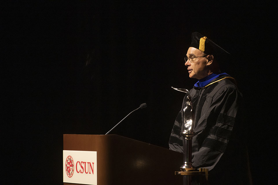 CSUN Faculty Senate President Michael Neubauer, in graduation regalia, speaks at a CSUN podium in a dark room.