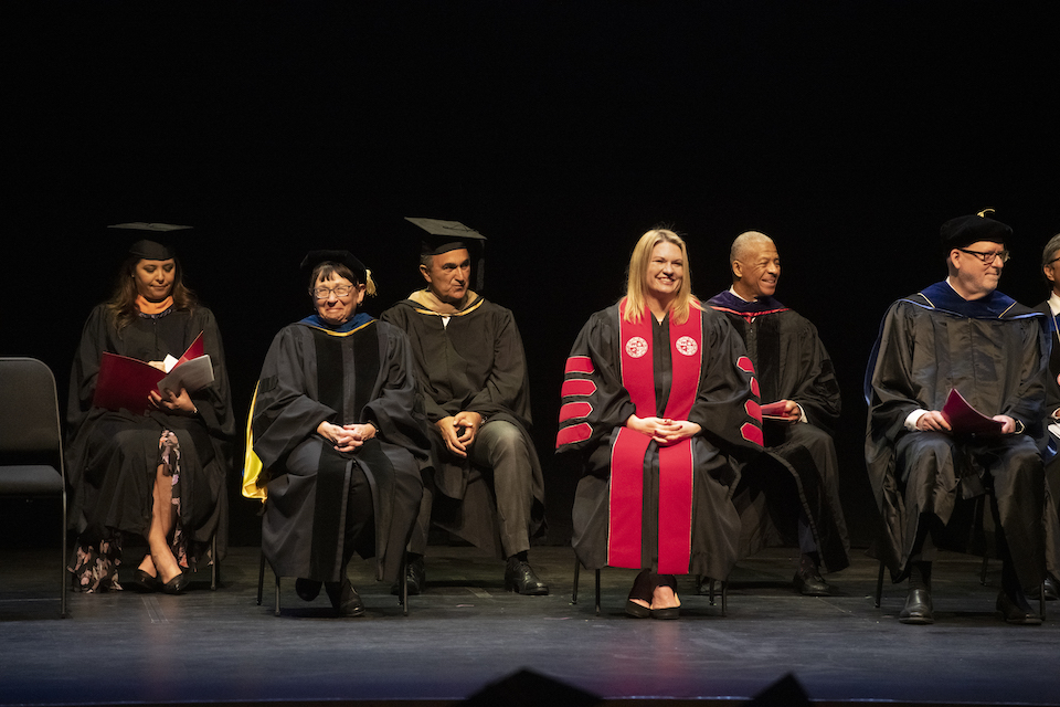 CSUN President Erika D. Beck in black academic robe with red sash with other academics in black robes on black floored stage